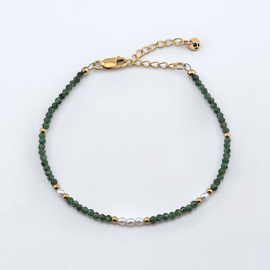 Top view of a dainty faceted emeralds and accent rice pearls high quality AAA  beaded bracelet with 14K gold filled accent beads, clasp, reverie blossom brand logo tag and extender chain laying flat on a plain off white or light gray background.