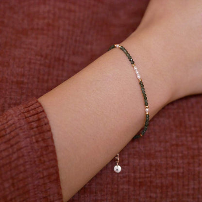 Close-up of a wrist wearing dainty  faceted emeralds and accent rice pearls high quality AAA beaded bracelet, with 14K gold filled accent beads. The background is a person wearing a brown long sleeve sweater.