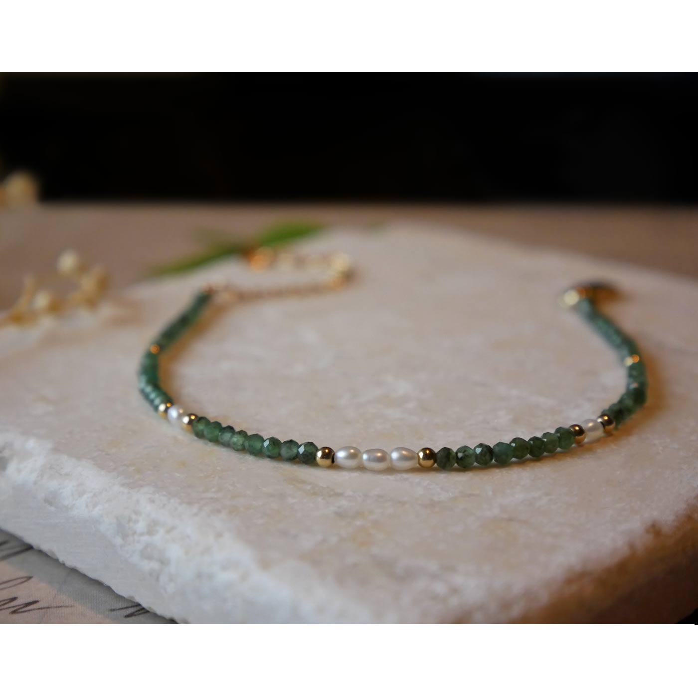 Front view of dainty faceted emeralds and accent rice pearls high quality AAA beaded bracelet, with 14K gold filled accent beads. It is laying flat on a stone surface stylized with a background of vintage light pink beige paper underneath with old cursive writing, and dried beige baby breath flowers.