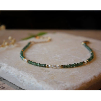 Front view of dainty faceted emeralds and accent rice pearls high quality AAA beaded bracelet, with 14K gold filled accent beads. It is laying flat on a stone surface stylized with a background of vintage light pink beige paper underneath with old cursive writing, and dried beige baby breath flowers.