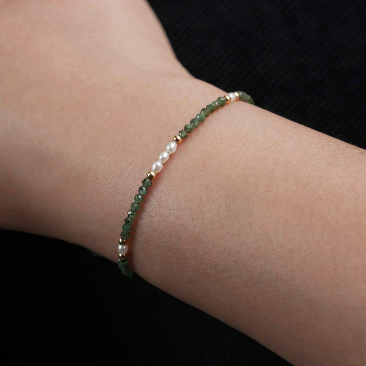 Close-up of a wrist wearing a dainty beaded bracelet with faceted emeralds, rice pearls, and 14K gold filled accents. The beads are high quality AAA, the background is dark black.