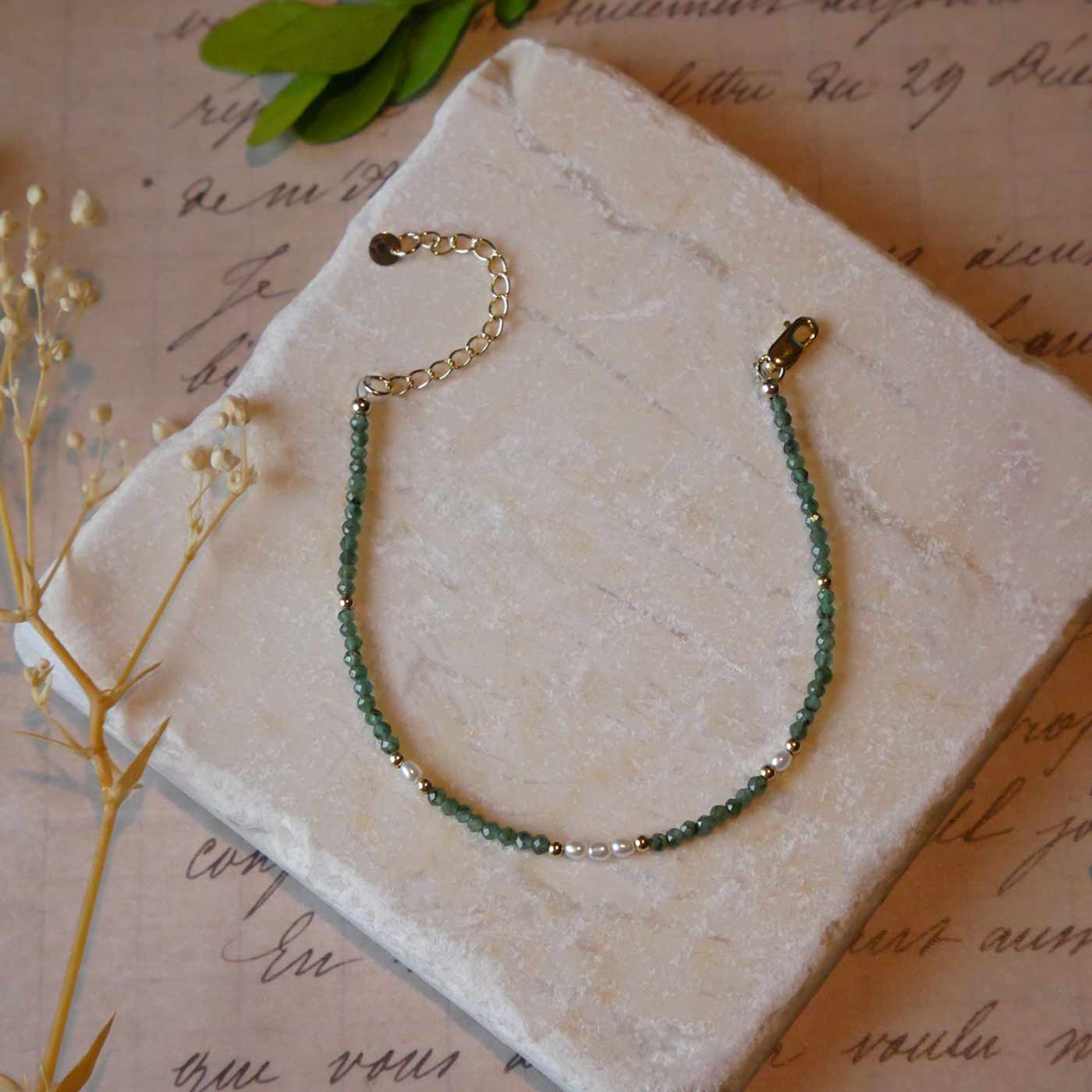 Top view of a dainty beaded bracelet with AAA quality faceted natural emeralds, rice pearls and 14K gold filled accent beads. It is laying flat on a stone surface stylized with a background of vintage light pink beige paper underneath with old cursive writing, dried beige baby breath flowers and green leaves.