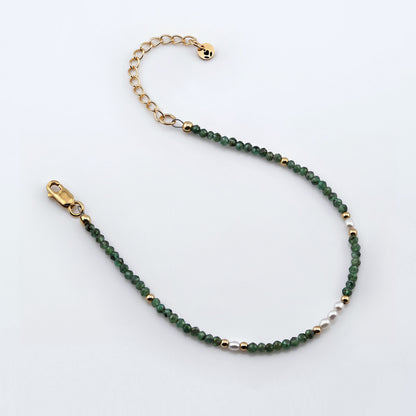 Top view of a dainty beaded bracelet with AAA quality faceted natural emeralds, rice pearls, 14K gold filled accent beads, clasp, reverie blossom brand logo tag and extender chain laying flat on a plain off white or light gray background.