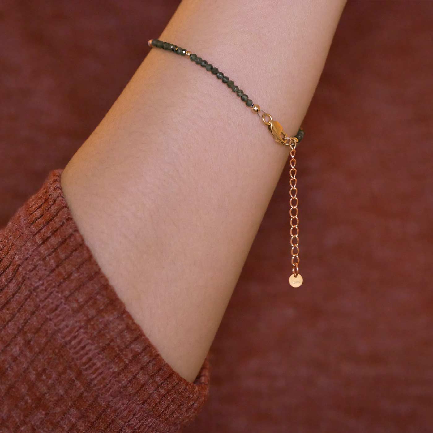 Close up of a person wearing a brown long sleeve shirt, wearing on their wrist a dainty beaded bracelet with AAA quality faceted natural emeralds, rice pearls and 14K gold filled accent beads, clasp, and extender chain hanging.
