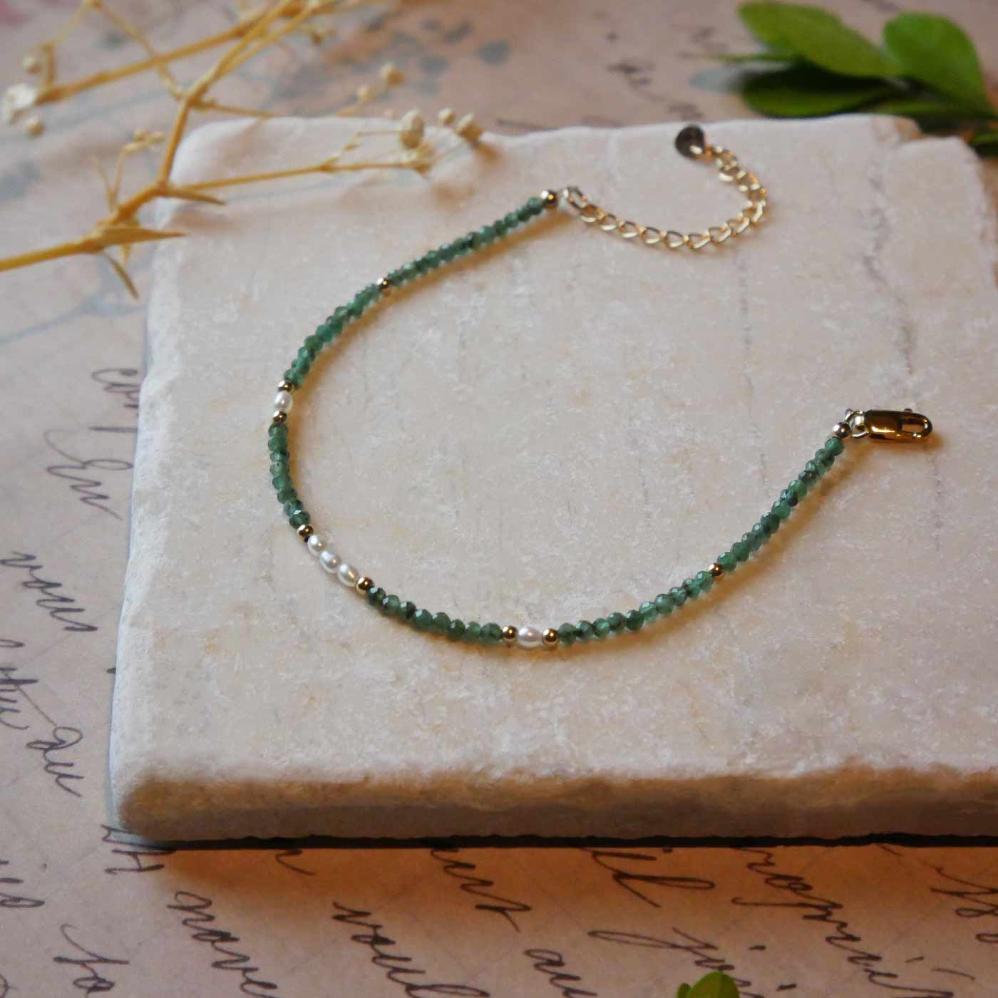 Top 3/4 view of a dainty beaded bracelet with AAA quality faceted natural emeralds, rice pearls and 14K gold filled accent beads. It is laying flat on a stone surface stylized with a background of vintage light pink beige paper underneath with old cursive writing, dried beige baby breath flowers and green leaves.