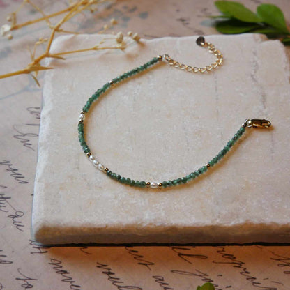 Top 3/4 view of a dainty beaded bracelet with AAA quality faceted natural emeralds, rice pearls and 14K gold filled accent beads. It is laying flat on a stone surface stylized with a background of vintage light pink beige paper underneath with old cursive writing, dried beige baby breath flowers and green leaves.