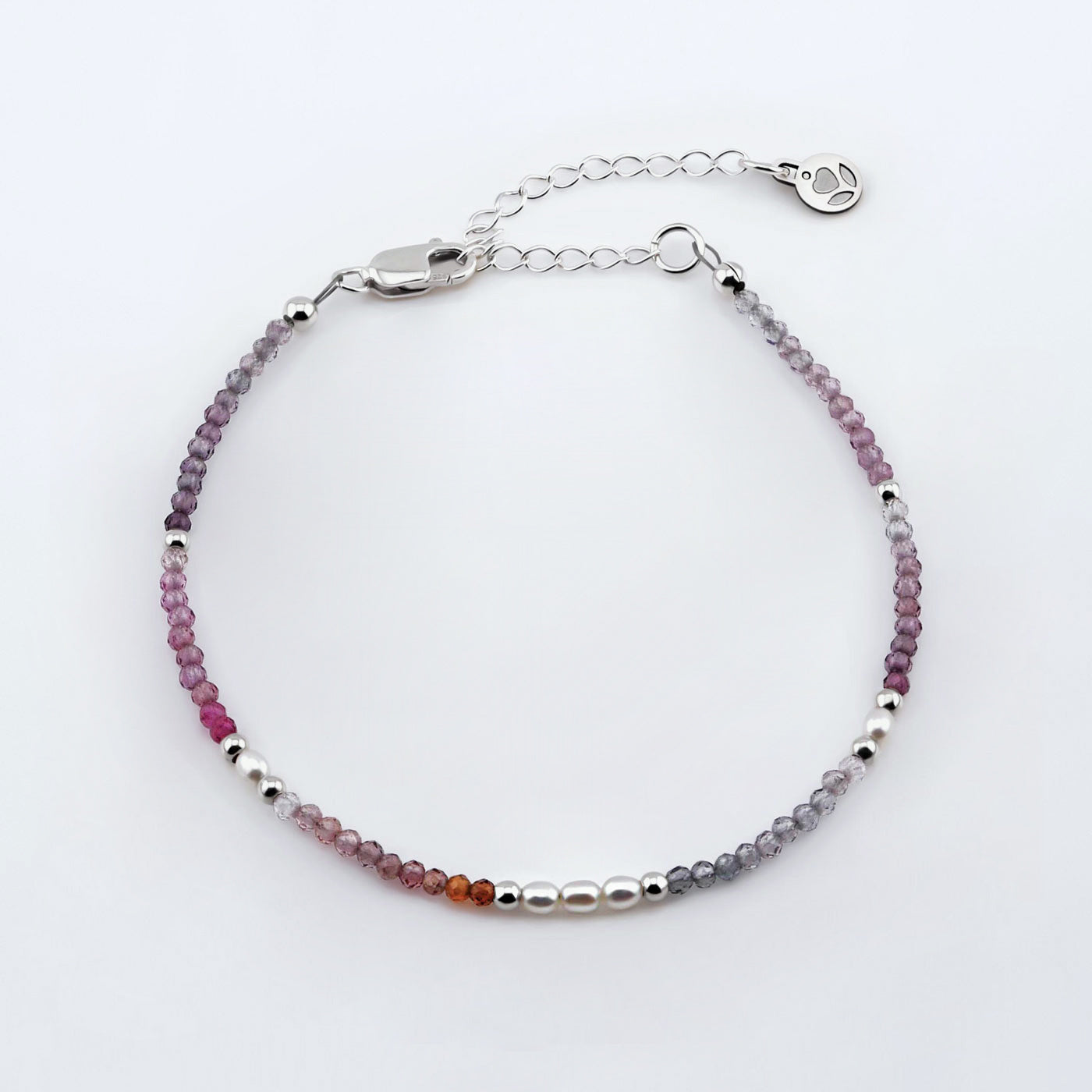 Top view of a dainty faceted colorful ombre spinel and accent rice pearls high quality AAA beaded bracelet with sterling silver accent beads, clasp, reverie blossom brand logo tag and extender chain laying flat on a plain off white or light gray background.