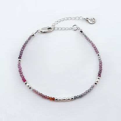 Top view of a dainty faceted colorful ombre spinel and accent rice pearls high quality AAA beaded bracelet with sterling silver accent beads, clasp, reverie blossom brand logo tag and extender chain laying flat on a plain off white or light gray background.