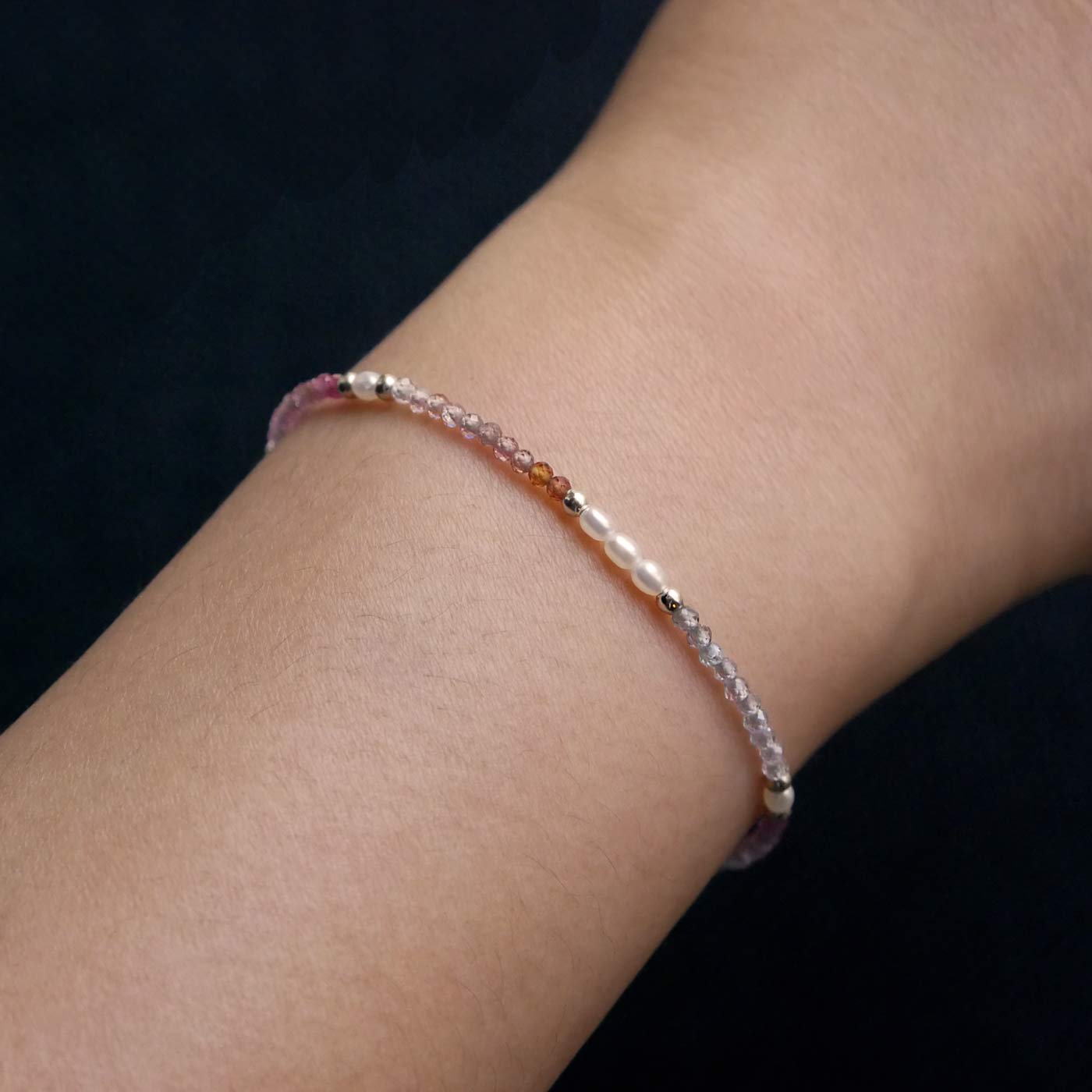 Close-up of a wrist wearing a dainty, delicate beaded bracelet with faceted colorful ombre spinel, rice pearls, and sterling silver accents. The beads are high quality AAA, the background is dark black.