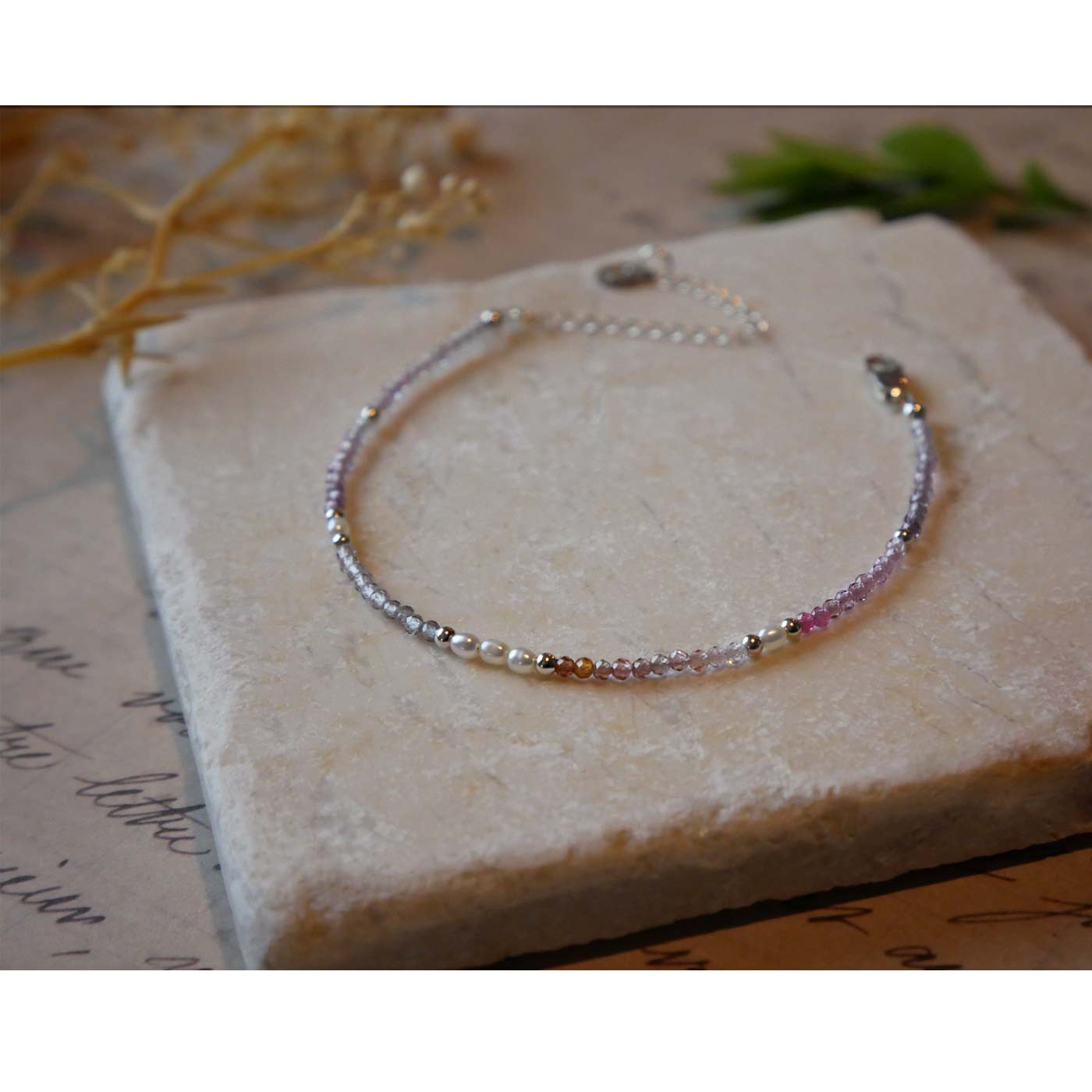 Top 3/4 view of a dainty beaded bracelet with AAA quality faceted natural colorful ombre spinel, rice pearls and sterling silver accent beads. It is laying flat on a stone surface stylized with a background of vintage light pink beige paper underneath with old cursive writing, dried beige baby breath flowers and green leaves.