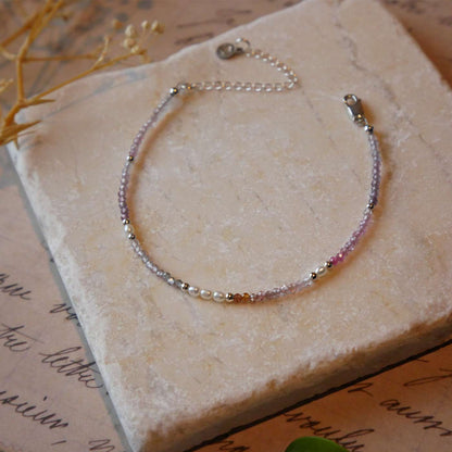Top 3/4 view of a dainty beaded bracelet with AAA quality faceted natural ombre colorful spinel, rice pearls and sterling silver accent beads. It is laying flat on a stone surface stylized with a background of vintage light pink beige paper underneath with old cursive writing, dried beige baby breath flower twigs and a green leaf.