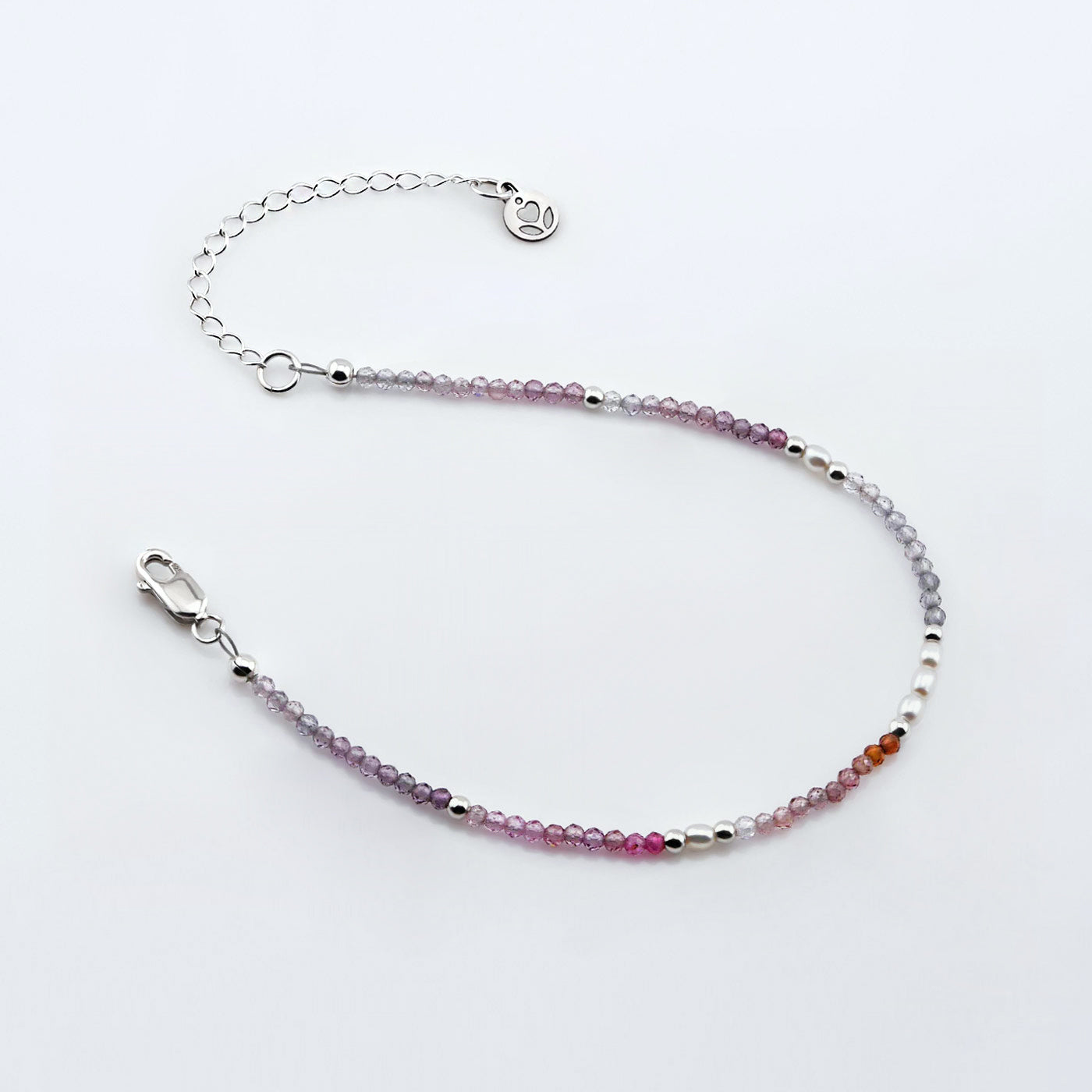 Top view of a dainty, delicate beaded bracelet with AAA quality faceted natural colorful ombre spinel, rice pearls, sterling silver accent beads, clasp, reverie blossom brand logo tag and extender chain laying flat on a plain off white or light gray background.