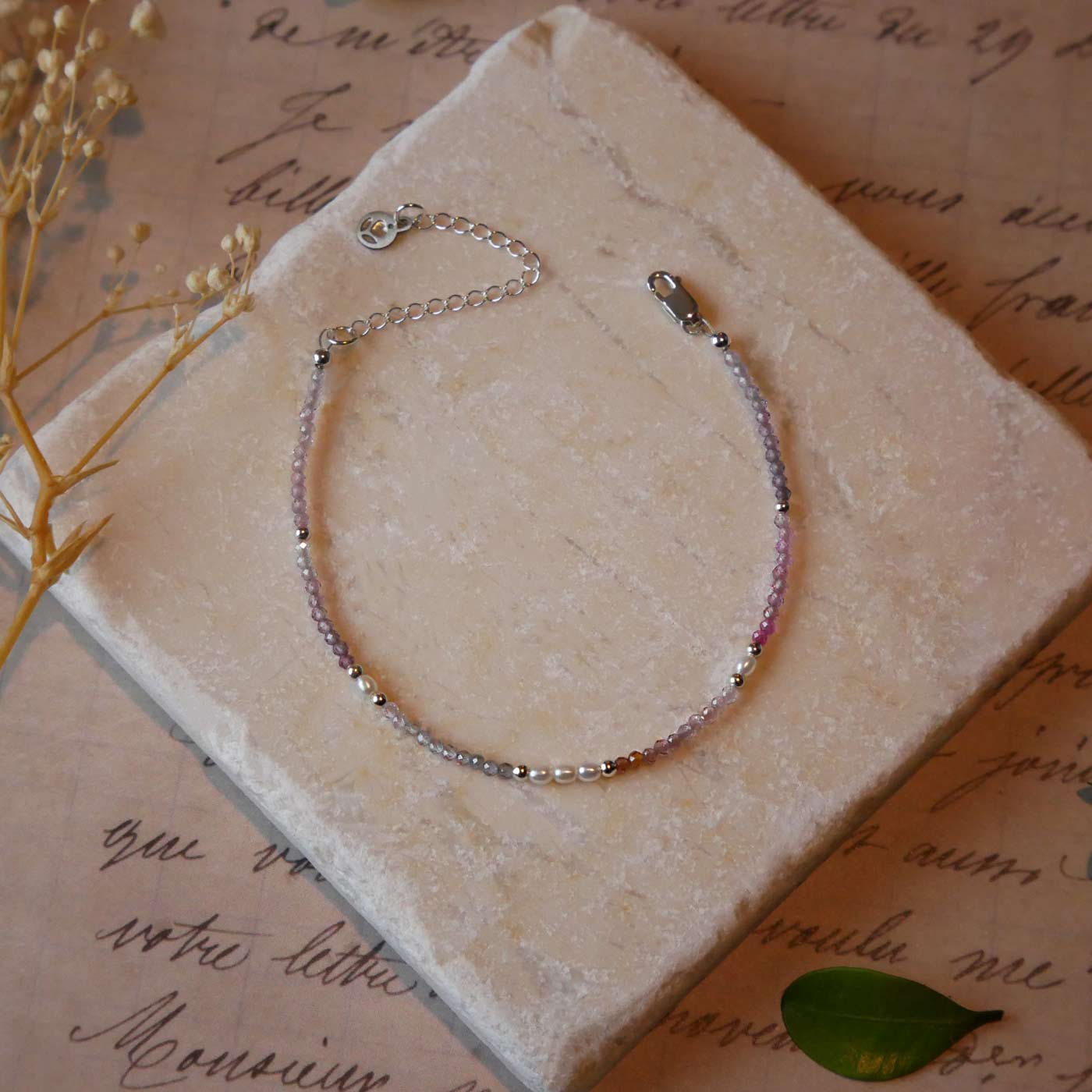 Top view of a dainty beaded bracelet with AAA quality faceted natural colorful ombre spinel, rice pearls and sterling silver accent beads. It is laying flat on a stone surface stylized with a background of vintage light pink beige paper underneath with old cursive writing, dried beige baby breath flowers and green leaves.