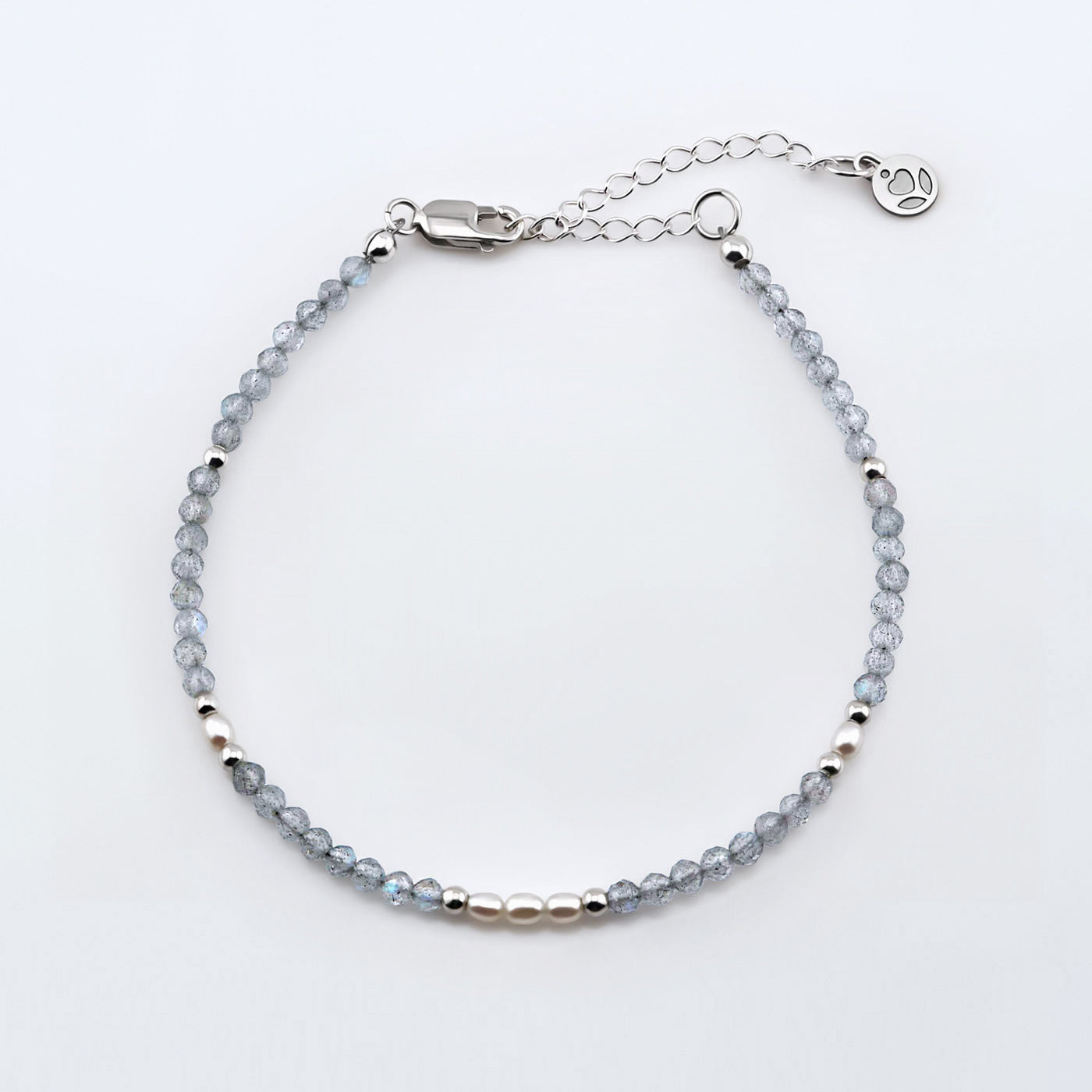 Top view of a dainty faceted labradorite and accent rice pearls high quality AAA  beaded bracelet with sterling silver accent beads, clasp, reverie blossom brand logo tag and extender chain laying flat on a plain off white or light gray background.