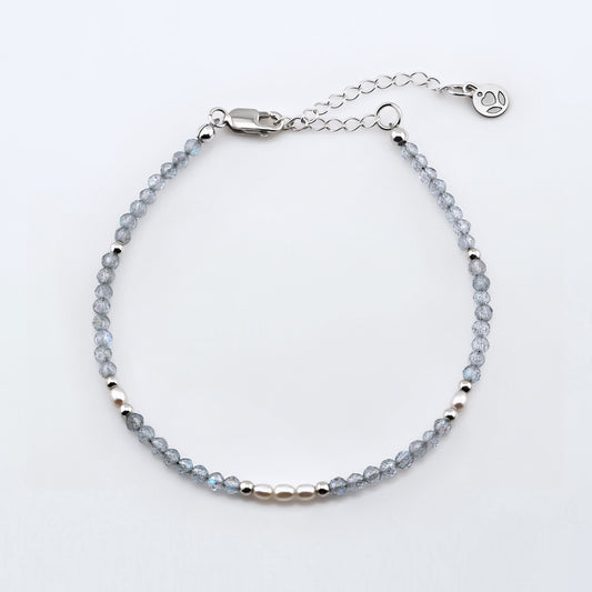 Top view of a dainty faceted labradorite and accent rice pearls high quality AAA  beaded bracelet with sterling silver accent beads, clasp, reverie blossom brand logo tag and extender chain laying flat on a plain off white or light gray background.