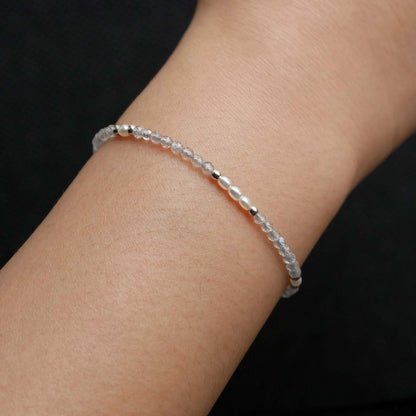 Close-up of a wrist wearing a dainty beaded bracelet with faceted labradorites, rice pearls, and sterling silver accents. The beads are high quality AAA, the background is dark black.