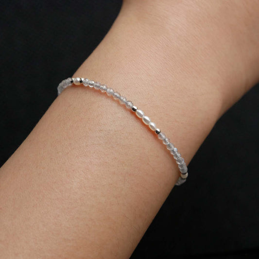 Close-up of a wrist wearing a dainty beaded bracelet with faceted labradorites, rice pearls, and sterling silver accents. The beads are high quality AAA, the background is dark black.