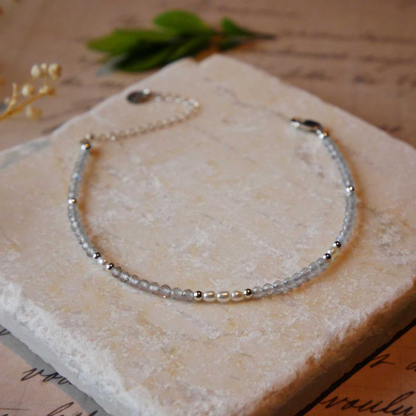 Front view of dainty faceted labradorite and accent rice pearls high quality AAA beaded bracelet, with sterling silver accent beads. It is laying flat on a stone surface stylized with a background of vintage light pink beige paper underneath with old cursive writing, and dried beige baby breath flowers.