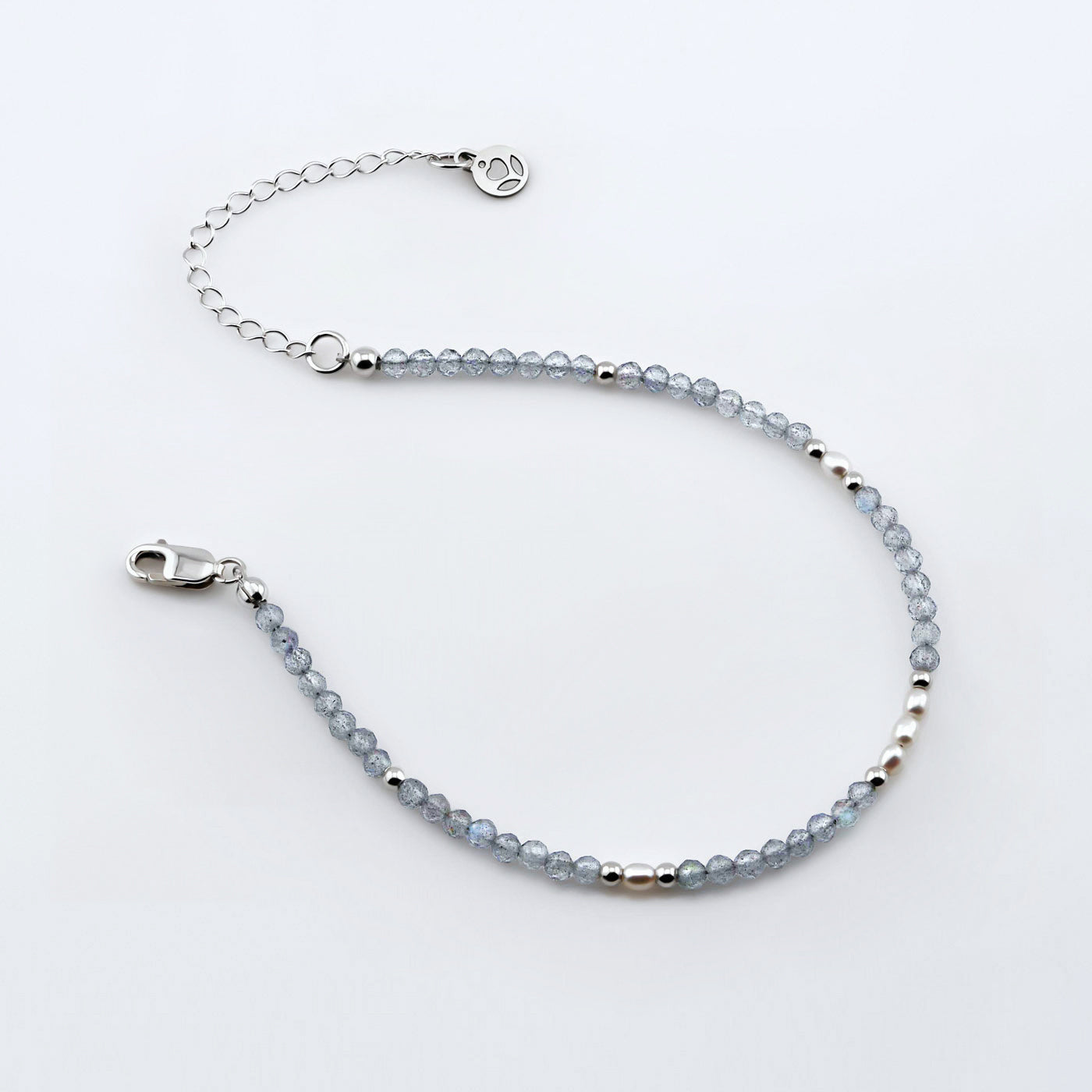 Top view of a dainty beaded bracelet with AAA quality faceted natural labradorites, rice pearls, sterling silver accent beads, clasp, reverie blossom brand logo tag and extender chain laying flat on a plain off white or light gray background.