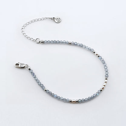 Top view of a dainty beaded bracelet with AAA quality faceted natural labradorites, rice pearls, sterling silver accent beads, clasp, reverie blossom brand logo tag and extender chain laying flat on a plain off white or light gray background.