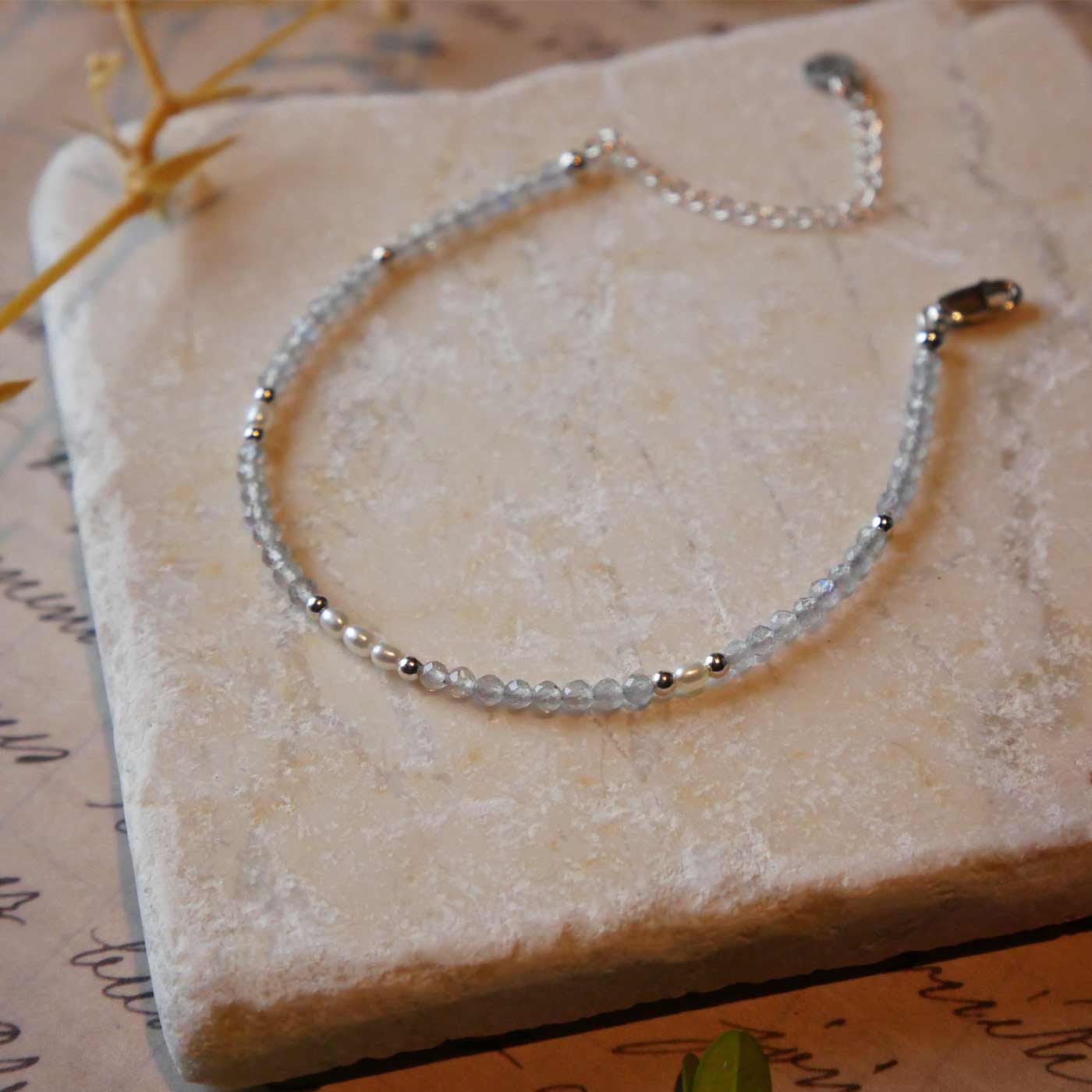 Top 3/4 view of a dainty beaded bracelet with AAA quality faceted natural labradorites, rice pearls and sterling silver accent beads. It is laying flat on a stone surface stylized with a background of vintage light pink beige paper underneath with old cursive writing, and dried beige baby breath flowers.