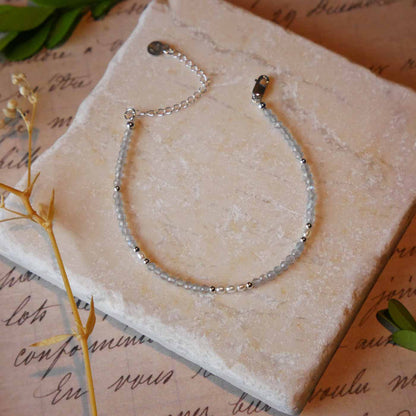 Top view of a dainty beaded bracelet with AAA quality faceted natural labradorites, rice pearls and sterling silver accent beads. It is laying flat on a stone surface stylized with a background of vintage light pink beige paper underneath with old cursive writing, dried beige baby breath flowers and green leaves.