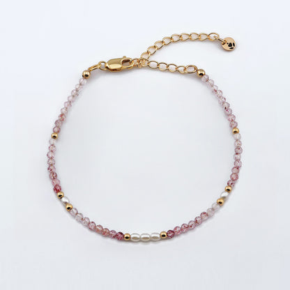 Top view of a dainty faceted strawberry quartz and accent rice pearls high quality AAA  beaded bracelet with 14K gold filled accent beads, clasp, reverie blossom brand logo tag and extender chain laying flat on a plain off white or light gray background.
