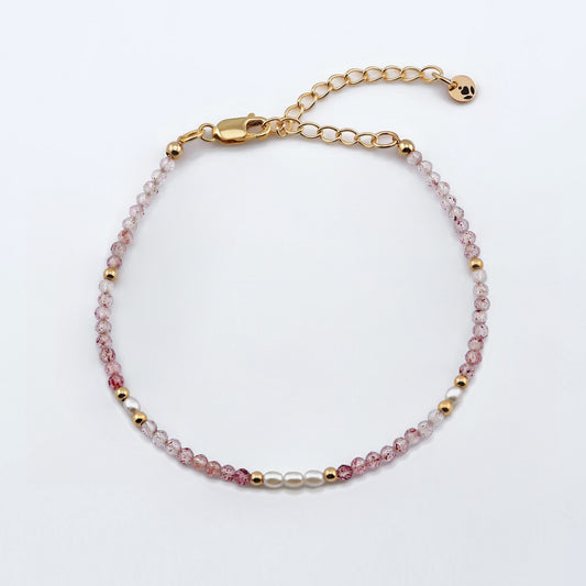 Top view of a dainty faceted strawberry quartz and accent rice pearls high quality AAA  beaded bracelet with 14K gold filled accent beads, clasp, reverie blossom brand logo tag and extender chain laying flat on a plain off white or light gray background.