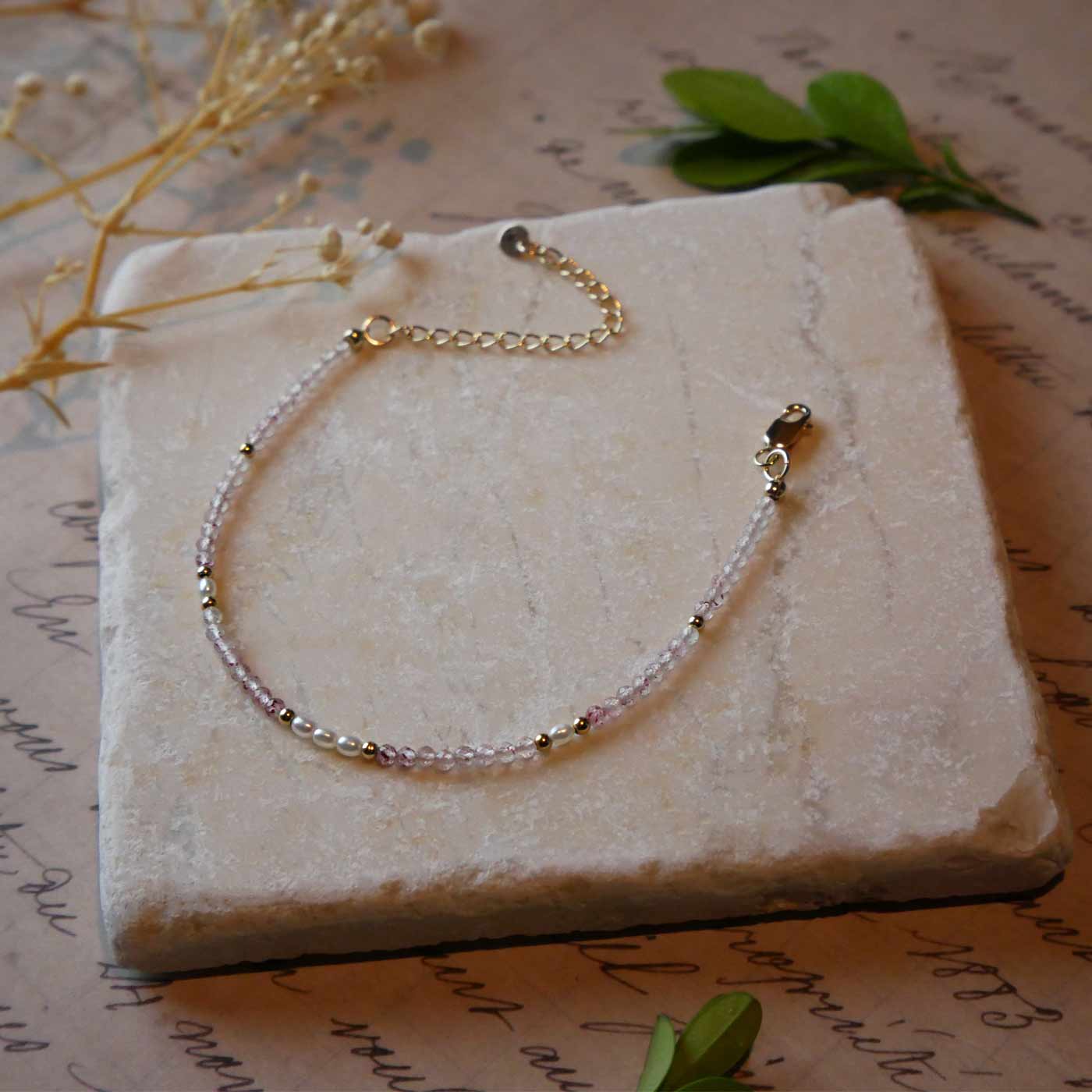 Top 3/4 view of a dainty beaded bracelet with AAA quality faceted natural strawberry quartz, rice pearls and 14K gold filled accent beads. It is laying flat on a stone surface stylized with a background of vintage light pink beige paper underneath with old cursive writing, dried beige baby breath flowers and green leaves.