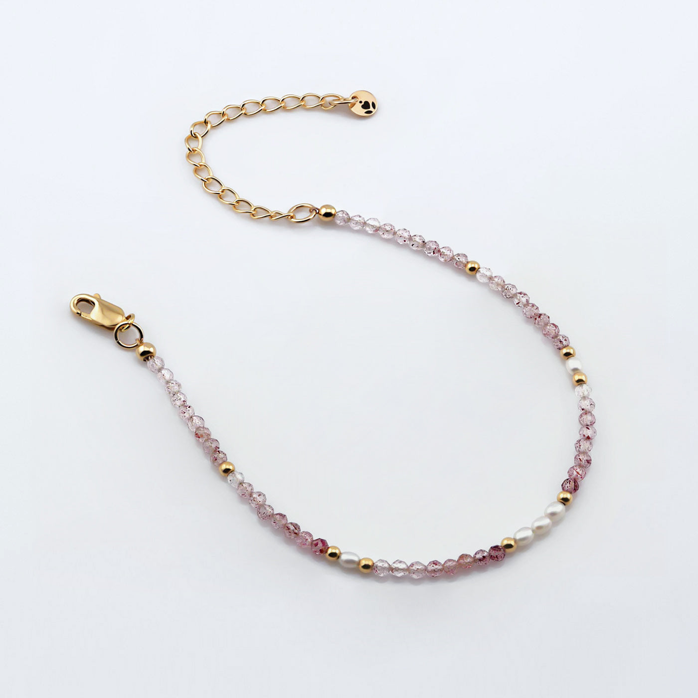 Top view of a dainty beaded bracelet with AAA quality faceted natural strawberry quartz, rice pearls, 14K gold filled accent beads, clasp, reverie blossom brand logo tag and extender chain laying flat on a plain off white or light gray background.