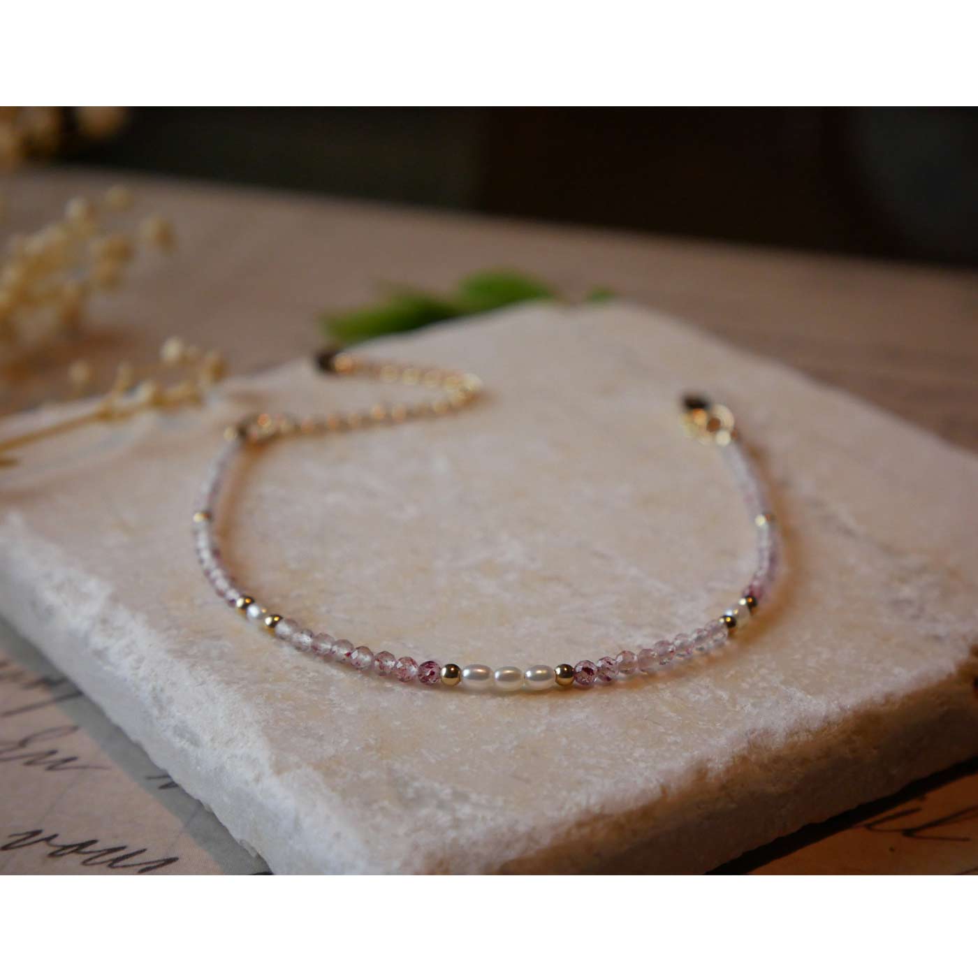 Front view of dainty faceted strawberry quartz and accent rice pearls high quality AAA beaded bracelet, with 14K gold filled accent beads. It is laying flat on a stone surface stylized with a background of vintage light pink beige paper underneath with old cursive writing, and dried beige baby breath flowers.