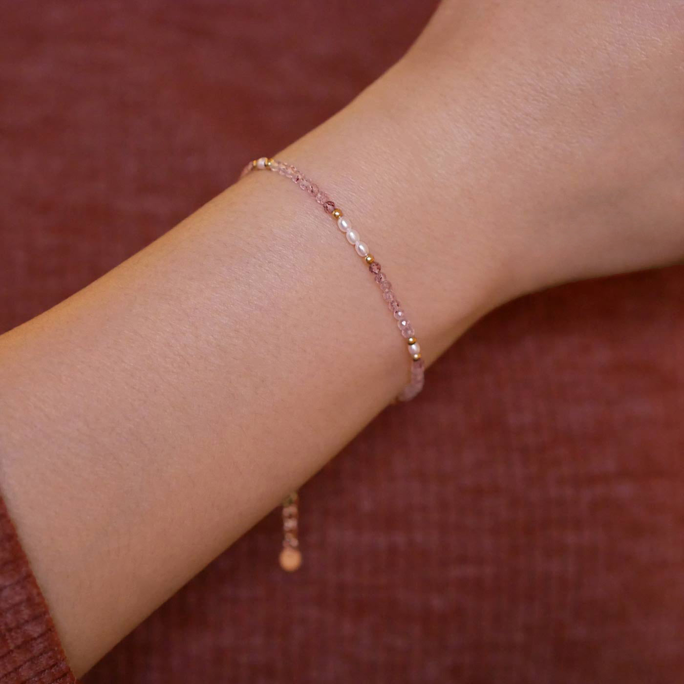 Close-up of a wrist wearing dainty, delicate faceted strawberry quartz and accent rice pearls high quality AAA beaded bracelet, with 14K gold filled accent beads. The background is a person wearing a brown long sleeve sweater.