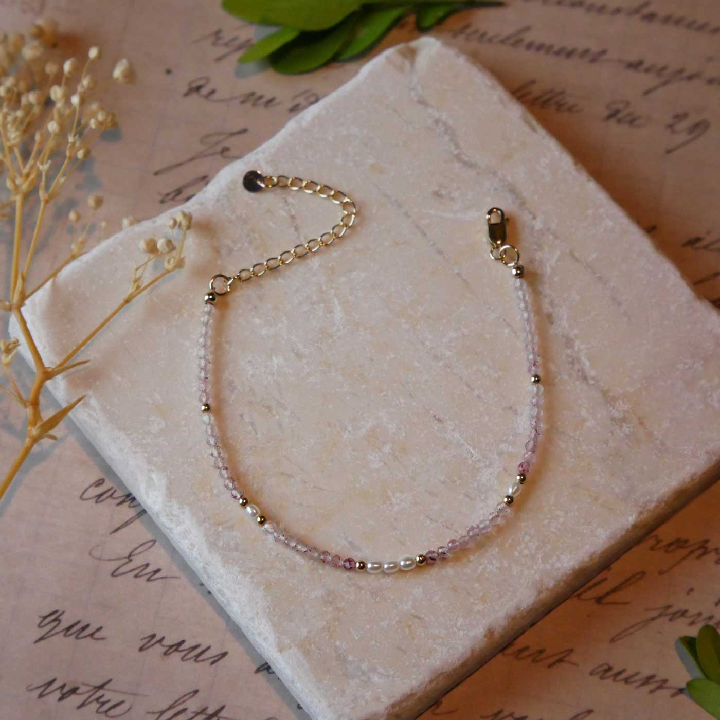 Top view of a dainty beaded bracelet with AAA quality faceted natural strawberry quartz, rice pearls and 14K gold filled accent beads. It is laying flat on a stone surface stylized with a background of vintage light pink beige paper underneath with old cursive writing, dried beige baby breath flowers and green leaves.