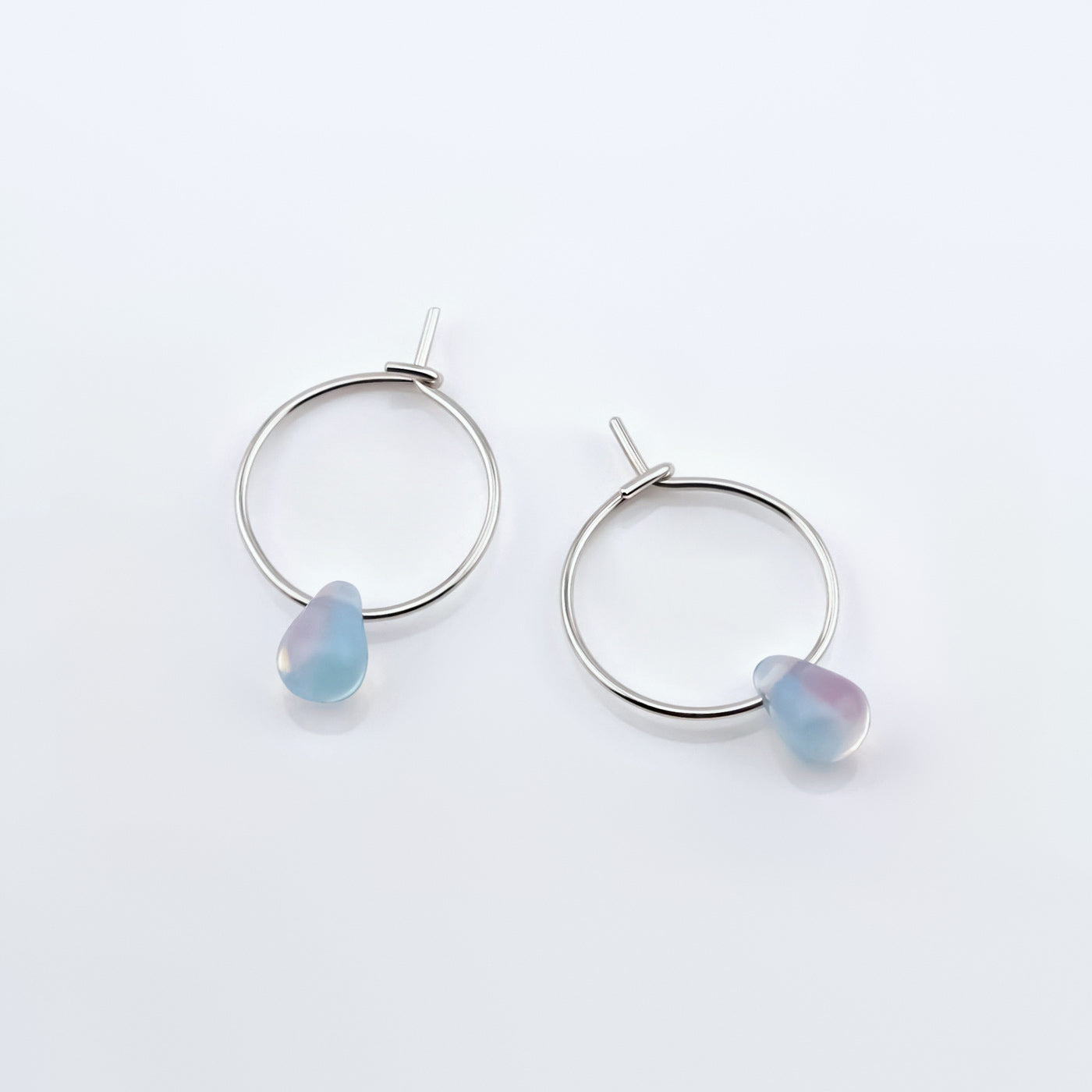 Top view of a pair of sterling silver Czech glass crystal milky blue pink colored drop wire hoop earrings, laying flat on a plain white background.
