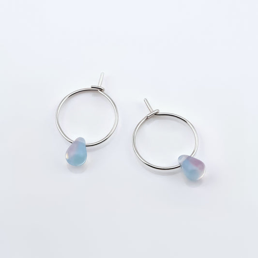 Top view of a pair of sterling silver Czech glass crystal milky blue pink colored drop wire hoop earrings, laying flat on a plain white background.