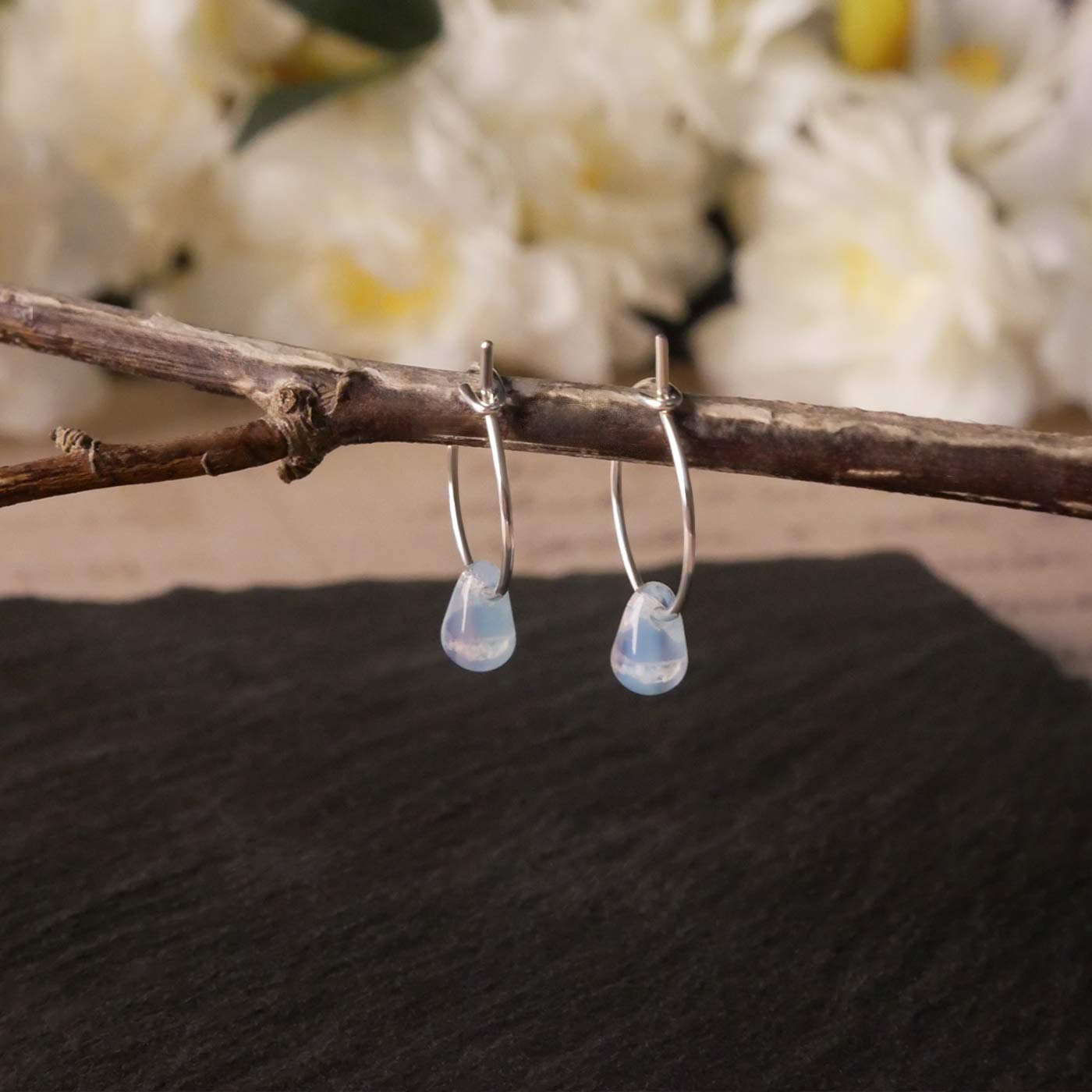 A pair of sterling silver Czech glass crystal milky blue pink colored drop wire hoop earrings, stylized by dangling on a branch over natural dark gray black stone surface, with blurred white flowers in the background.