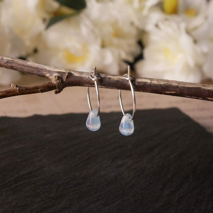 A pair of sterling silver Czech glass crystal milky blue pink colored drop wire hoop earrings, stylized by dangling on a branch over natural dark gray black stone surface, with blurred white flowers in the background.