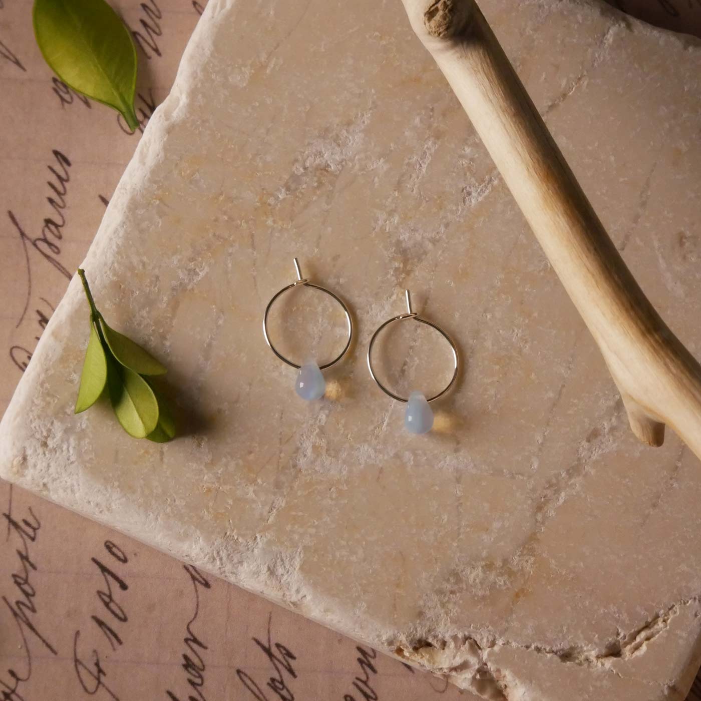 Top view with a pair of sterling silver Czech glass crystal, milky blue pink colored drop wire hoop earrings. It is stylized by laying flat on a natural stone surface, over vintage paper with cursive writing, and surrounded by green leaves with a branch in the foreground.
