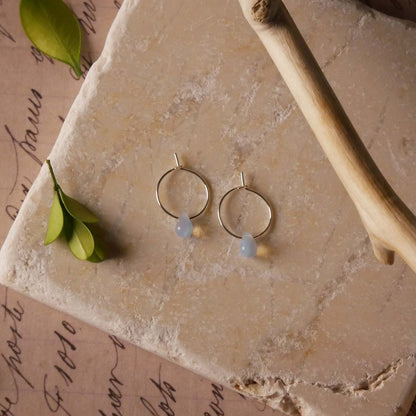 Top view with a pair of sterling silver Czech glass crystal, milky blue pink colored drop wire hoop earrings. It is stylized by laying flat on a natural stone surface, over vintage paper with cursive writing, and surrounded by green leaves with a branch in the foreground.