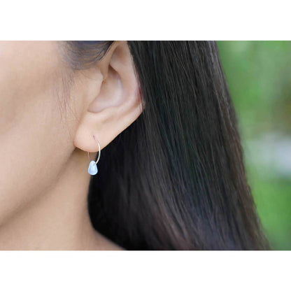 A close up of a sterling silver Czech glass crystal milky blue pink colored drop wire hoop earring, worn outside by a person with dark hair. The background is blurred with plants in the background.