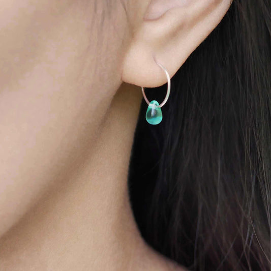 A close up of a sterling silver Czech glass crystal drop in a teal color on a wire hoop earring, worn outside by a person with dark hair.