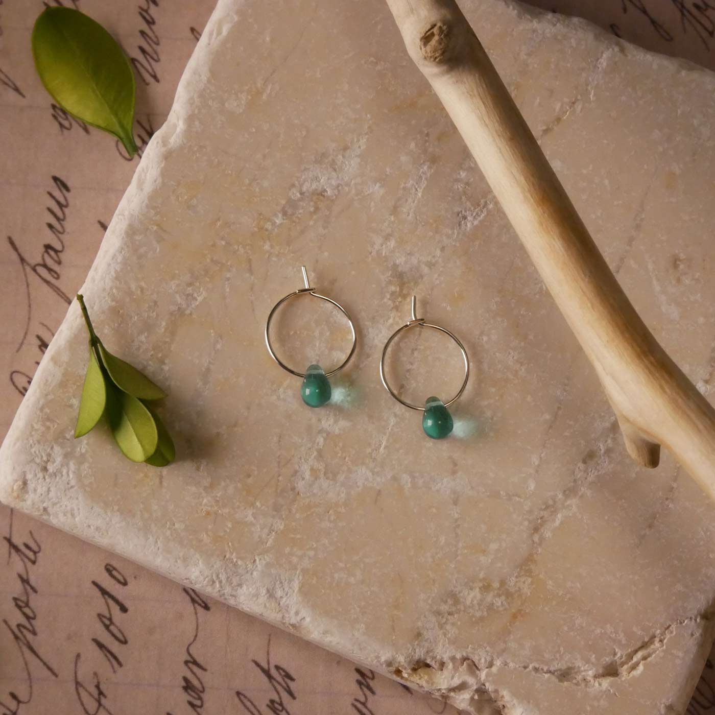 Top view with a pair of sterling silver Czech glass crystal, teal colored drop wire hoop earrings. It is stylized by laying flat on a natural stone surface, over vintage paper with cursive writing, and surrounded by green leaves with a branch in the foreground.