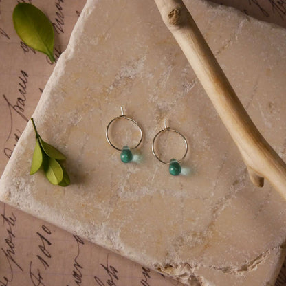 Top view with a pair of sterling silver Czech glass crystal, teal colored drop wire hoop earrings. It is stylized by laying flat on a natural stone surface, over vintage paper with cursive writing, and surrounded by green leaves with a branch in the foreground.