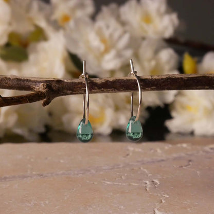 A pair of sterling silver Czech glass crystal teal colored drop wire hoop earrings, stylized by dangling on a branch over natural beige stone surface, with blurred white flowers in the background.