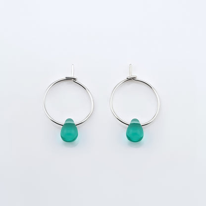 Top view of a pair of sterling silver Czech glass crystal teal colored drop wire hoop earrings, laying flat on a plain white background.
