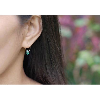 A close up of a sterling silver Czech glass crystal teal colored drop wire hoop earring, worn outside by a person with dark hair. The background is blurred with plants in the background.