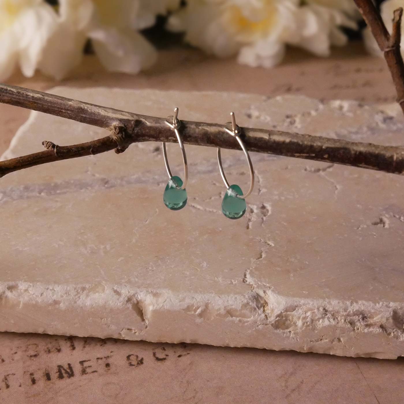 A pair of sterling silver Czech glass crystal teal colored drop wire hoop earrings, stylized by dangling on a branch over natural beige stone surface, vintage paper underneath, with blurred white flowers in the background.