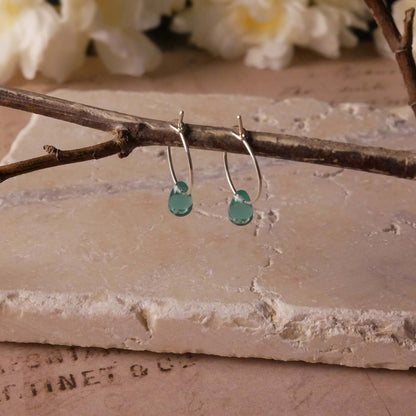 A pair of sterling silver Czech glass crystal teal colored drop wire hoop earrings, stylized by dangling on a branch over natural beige stone surface, vintage paper underneath, with blurred white flowers in the background.