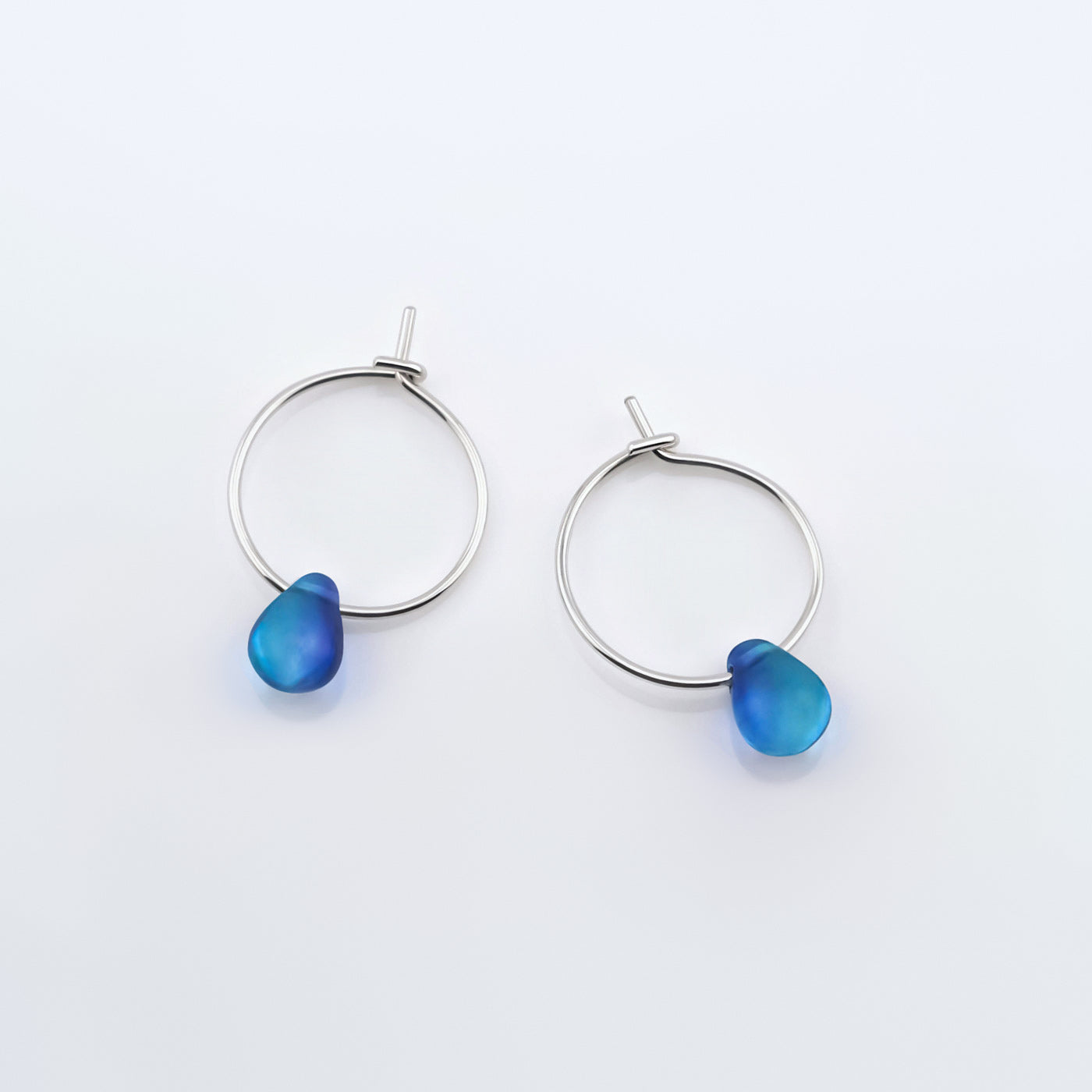 Top view of a pair of sterling silver Czech glass crystal blue violet teal colored drop wire hoop earrings, laying flat on a plain white background.