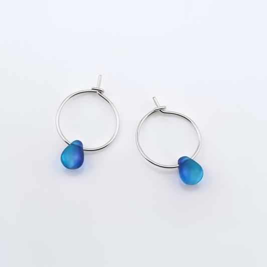 Top view of a pair of sterling silver Czech glass crystal blue violet teal colored drop wire hoop earrings, laying flat on a plain white background.